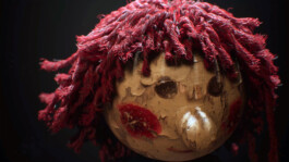Close-up of a heavily worn wooden doll’s head with red, frayed yarn hair, peeling paint on the face, red painted cheeks, and a dark background