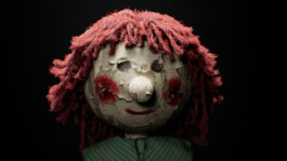Close-up of a heavily worn wooden doll’s head with red, frayed yarn hair, peeling paint on the face, red painted cheeks, and a dark background 